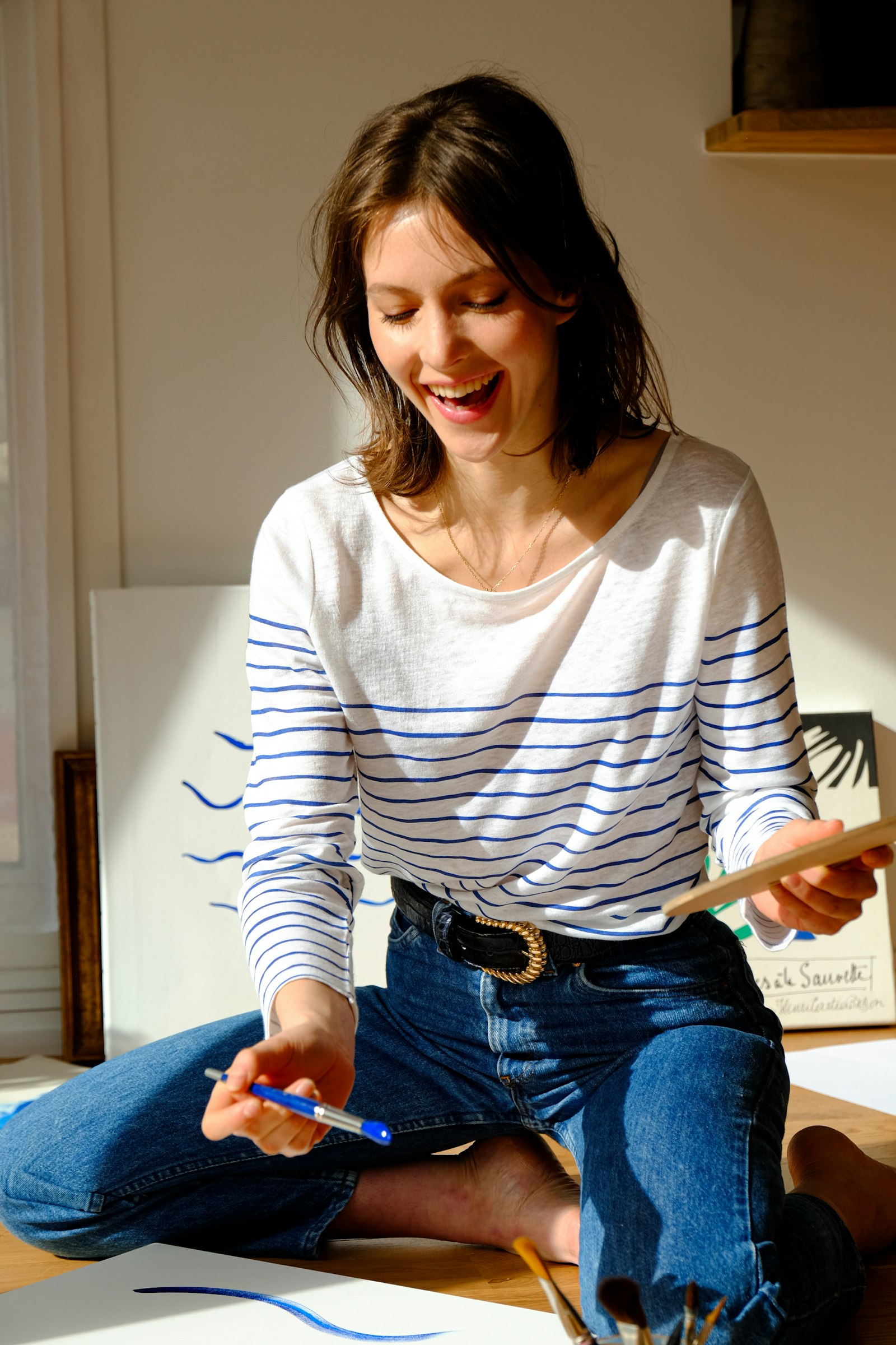 A printmaker in a blue-and-white striped top laughs while working cross-legged on the studio floor, canvases and a framed print behind her